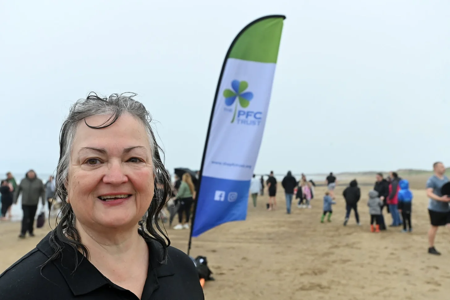 Woman smiling at camera on a beach with a PFC Trust banner behind her and groups of people gathered in the background.