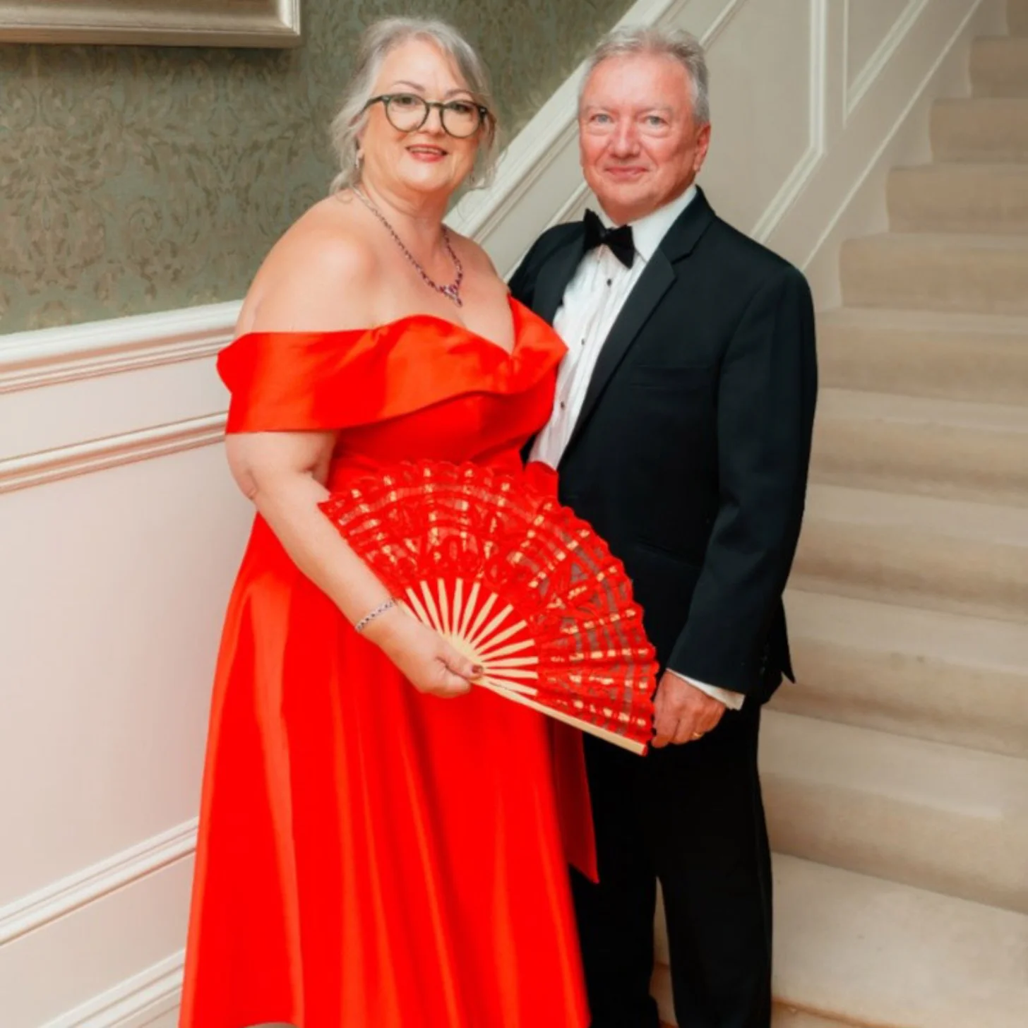 A woman in a red off-shoulder gown holding a red decorative fan stands next to a man in a black tuxedo with a bow tie in an elegant hallway.