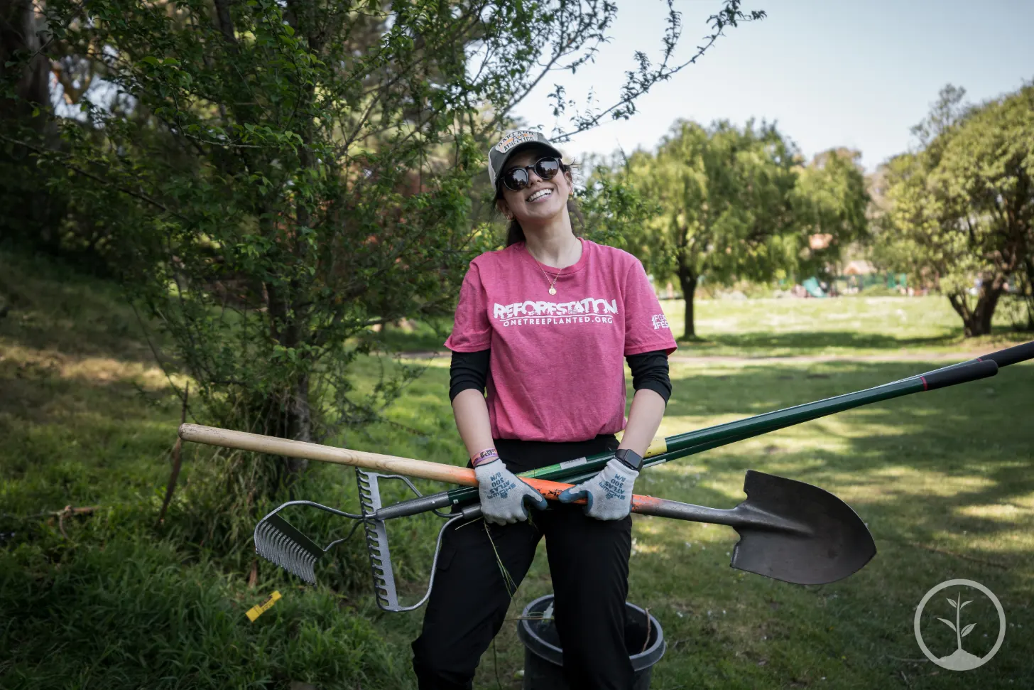 A person wearing a maroon shirt with "REFORESTATION" text holds gardening tools including a rake, shovel, and spade while standing in a park with trees in the background.
