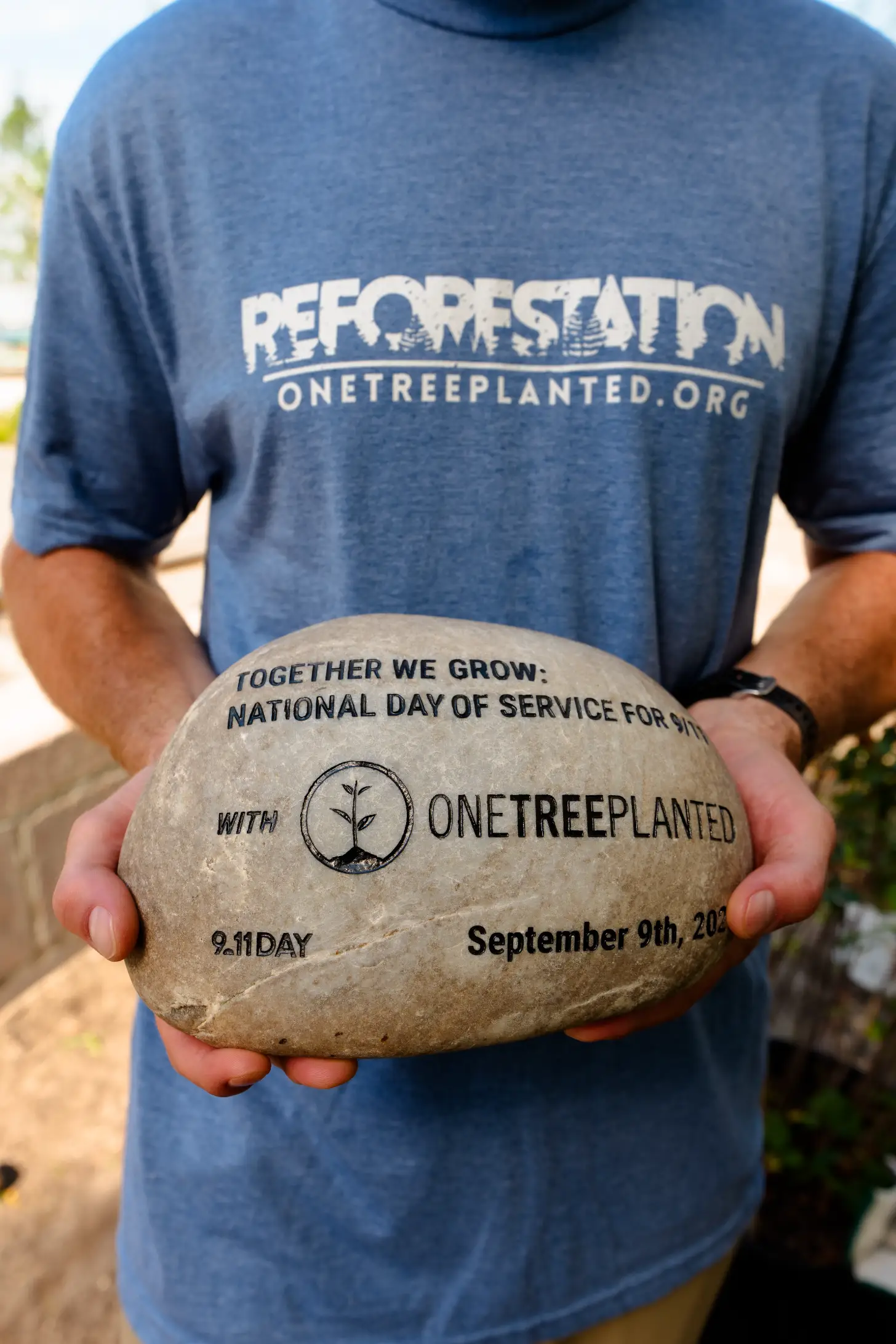 A person wearing a blue "Reforestation" shirt holds a stone engraved with "Together We Grow: National Day of Service for 9.11 Day September 9th, 2021 with OneTreePlanted" and a tree seedling logo.