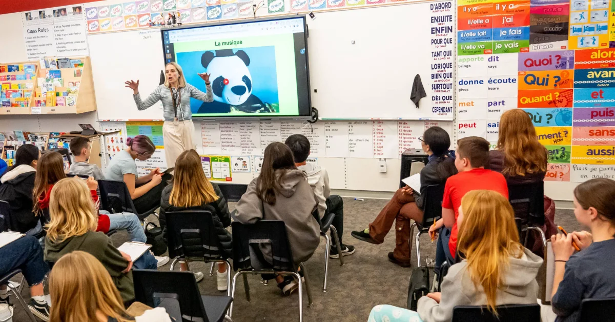 A French language teacher stands at the front of a classroom gesturing toward a interactive screen displaying a panda with text "La musique." Students seated on chairs face the screen, with classroom walls displaying French vocabulary posters and grammar charts.