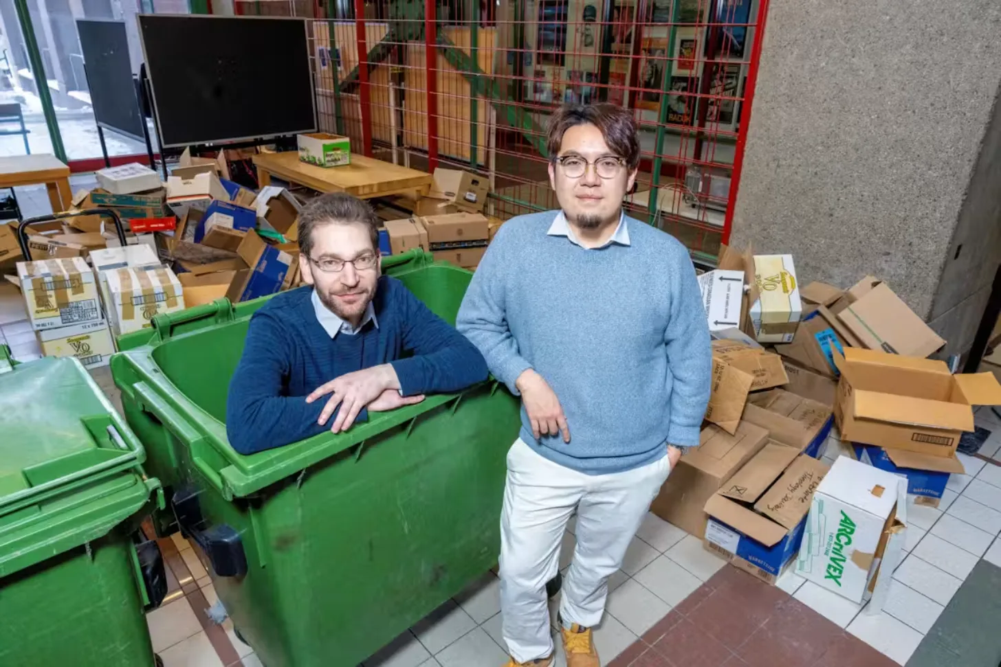 Two men stand in a cluttered room surrounded by cardboard boxes and green plastic bins. Shelves with colorful book spines line the wall behind them.