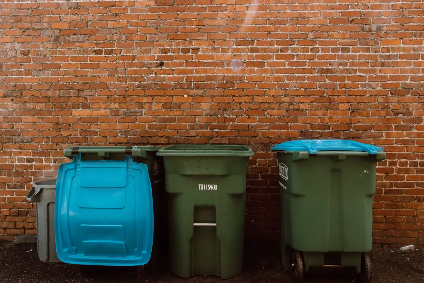 Three large waste bins lined against a brick wall: a blue bin on the left, two green bins in the center and right with blue lids. The center green bin displays the number T0115400.