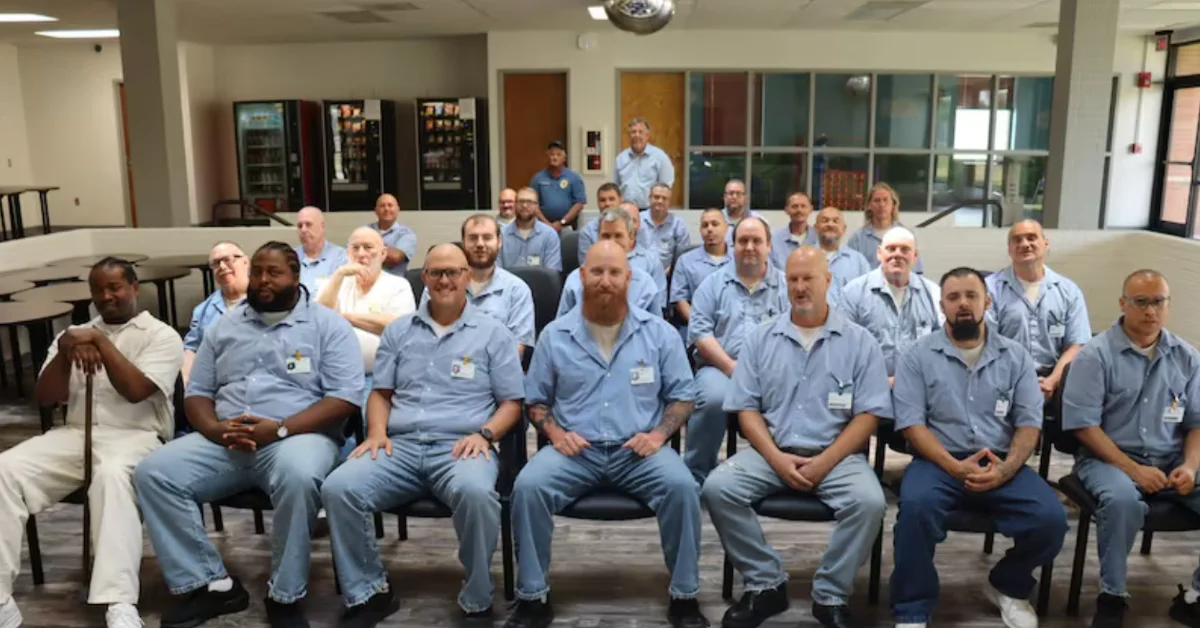 Group of men wearing light blue work shirts seated and standing in rows in an institutional cafeteria setting with vending machines and large windows visible in the background.