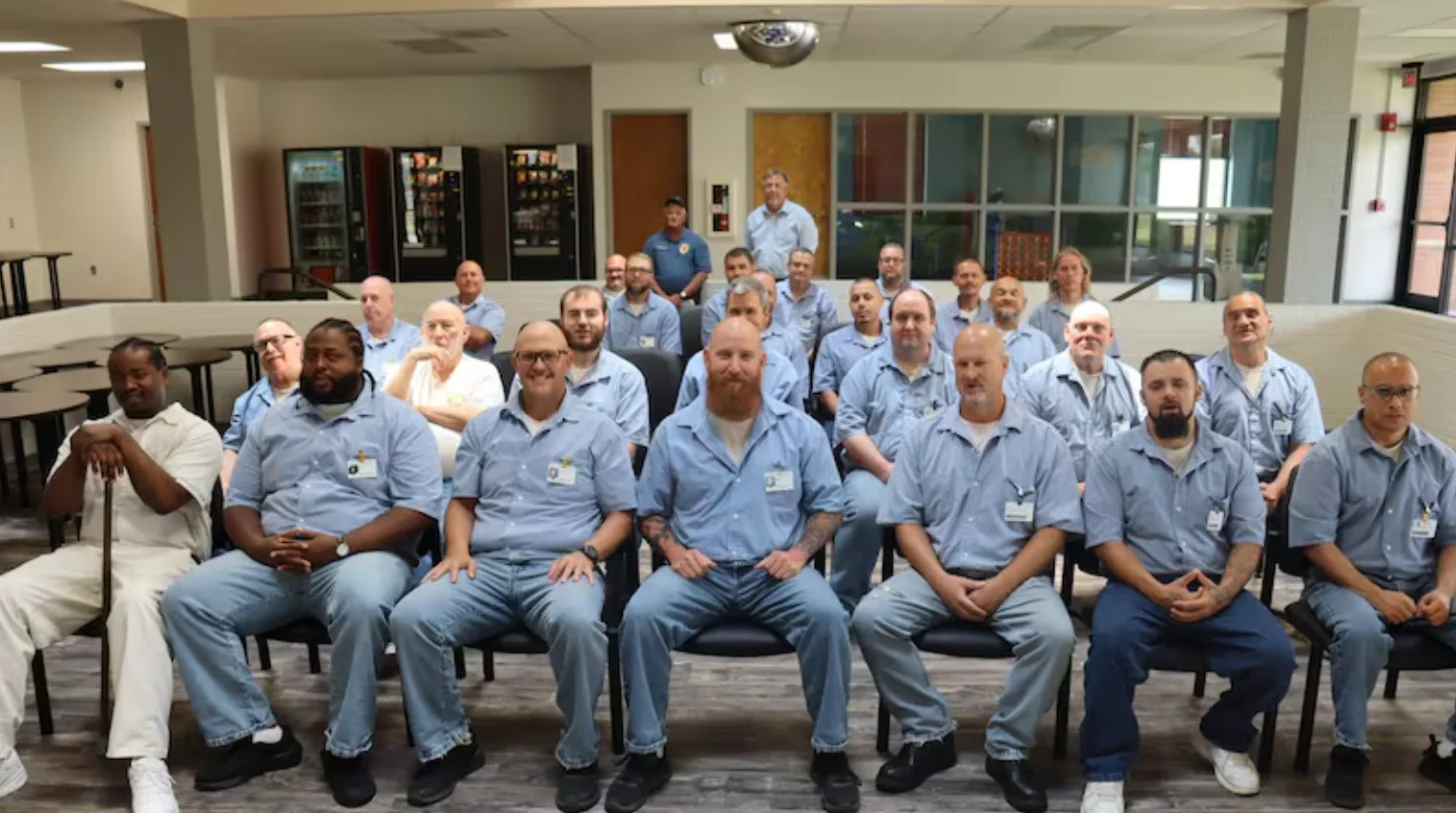 Large group of men wearing light blue work shirts seated and standing in a cafeteria or break room, with vending machines and windows visible in the background.