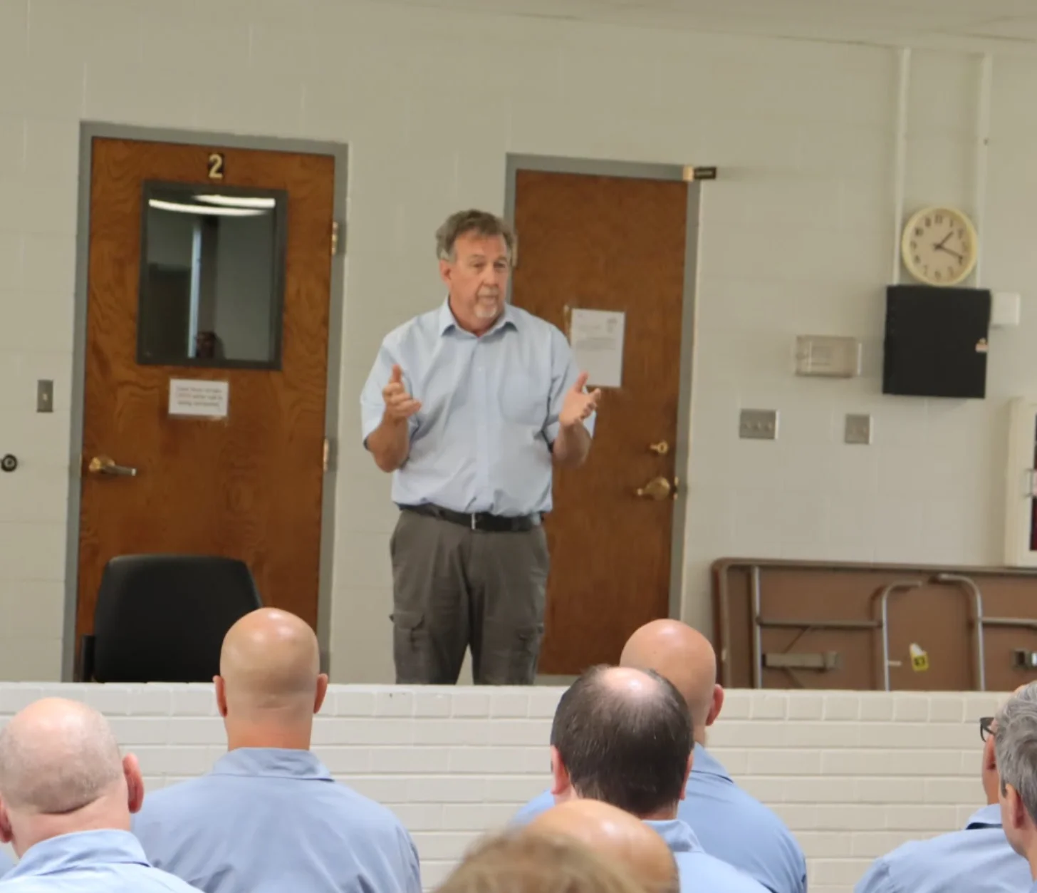A man in a light blue shirt and tan pants stands speaking with his hands raised in a room with brown doors marked with the number 2. Several people wearing blue shirts sit with their backs to the camera, listening.