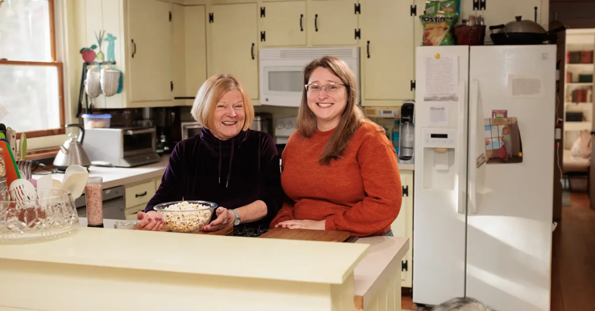 Two women stand at a kitchen counter, one wearing a dark purple top and the other wearing an orange sweater, both smiling at the camera. The woman on the left holds a bowl of popcorn.