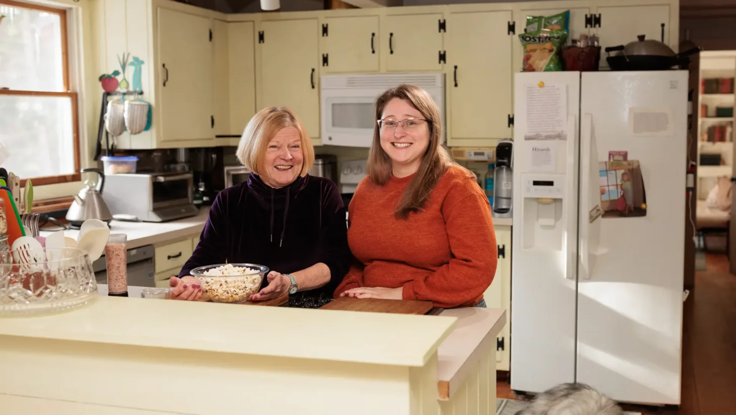 Two women stand at a kitchen counter holding a bowl of popcorn, both smiling at the camera. The kitchen behind them features cream-colored cabinets, a white refrigerator, and various countertop items.