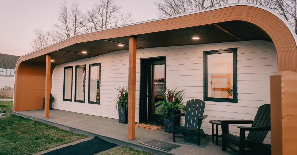 A modern curved wooden structure with white horizontal siding and wood columns features a covered porch with black-framed windows and doors. Two dark green Adirondack chairs and a small table sit on the concrete patio in front. Bare trees are visible in the background.