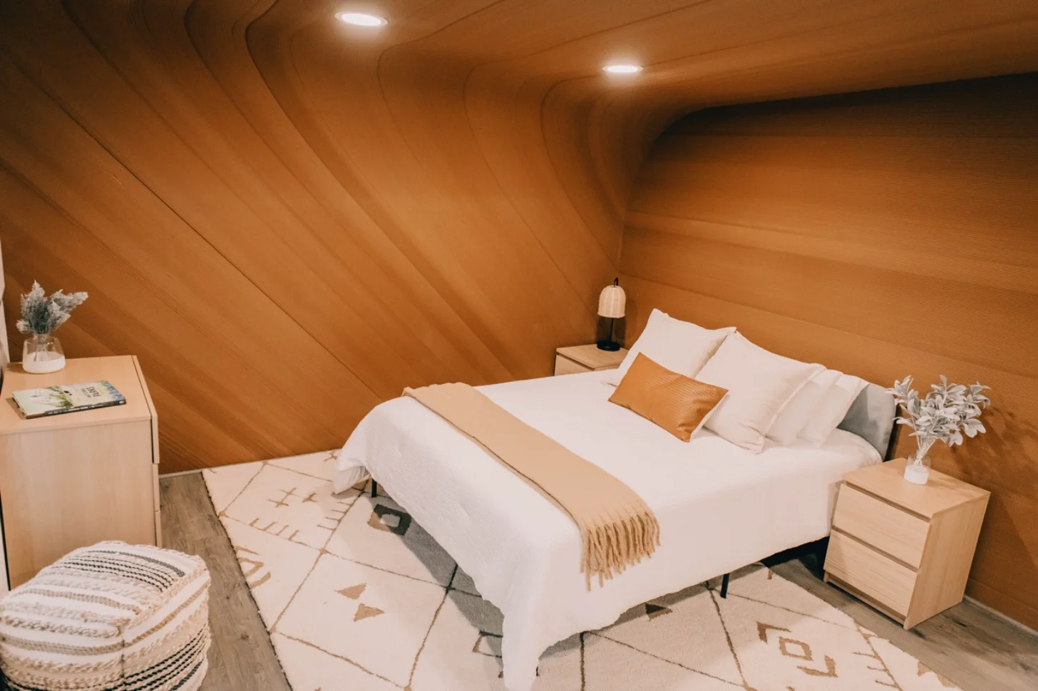 A bedroom with wood-paneled sloped walls features a bed with white linens, a tan throw blanket, and decorative pillows. Light wood nightstands flank the bed, one holding a pendant lamp and potted plant. A woven pouf sits on a geometric patterned rug in the foreground, with a wooden dresser and vase with dried flowers visible on the left.