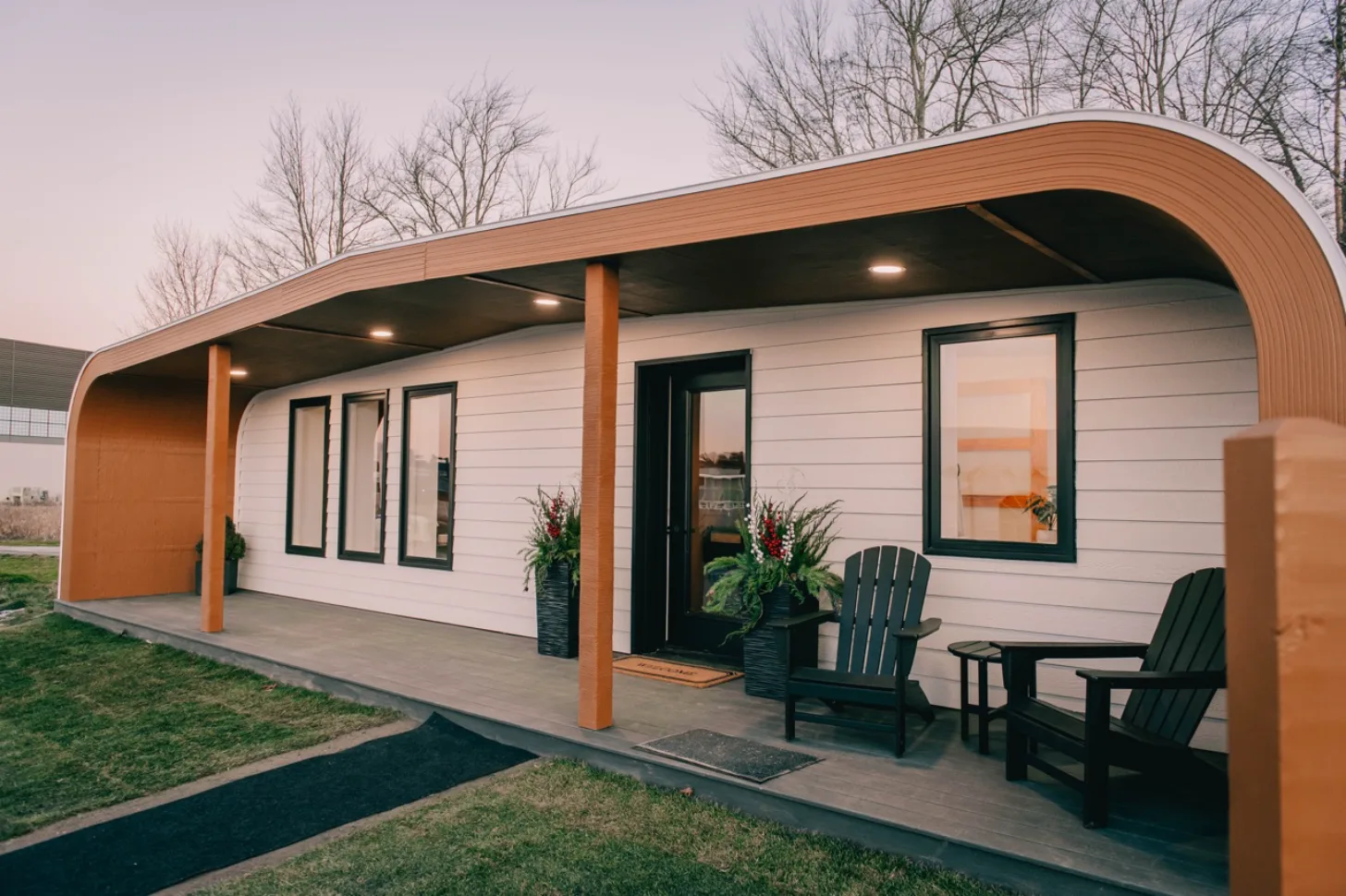 A modern residential structure with white horizontal siding and a curved orange-brown roof features a covered porch with wooden decking, orange support columns, and recessed lighting. Two dark green Adirondack chairs and a small table sit on the porch near potted plants and the entrance door. Bare trees are visible in the background.