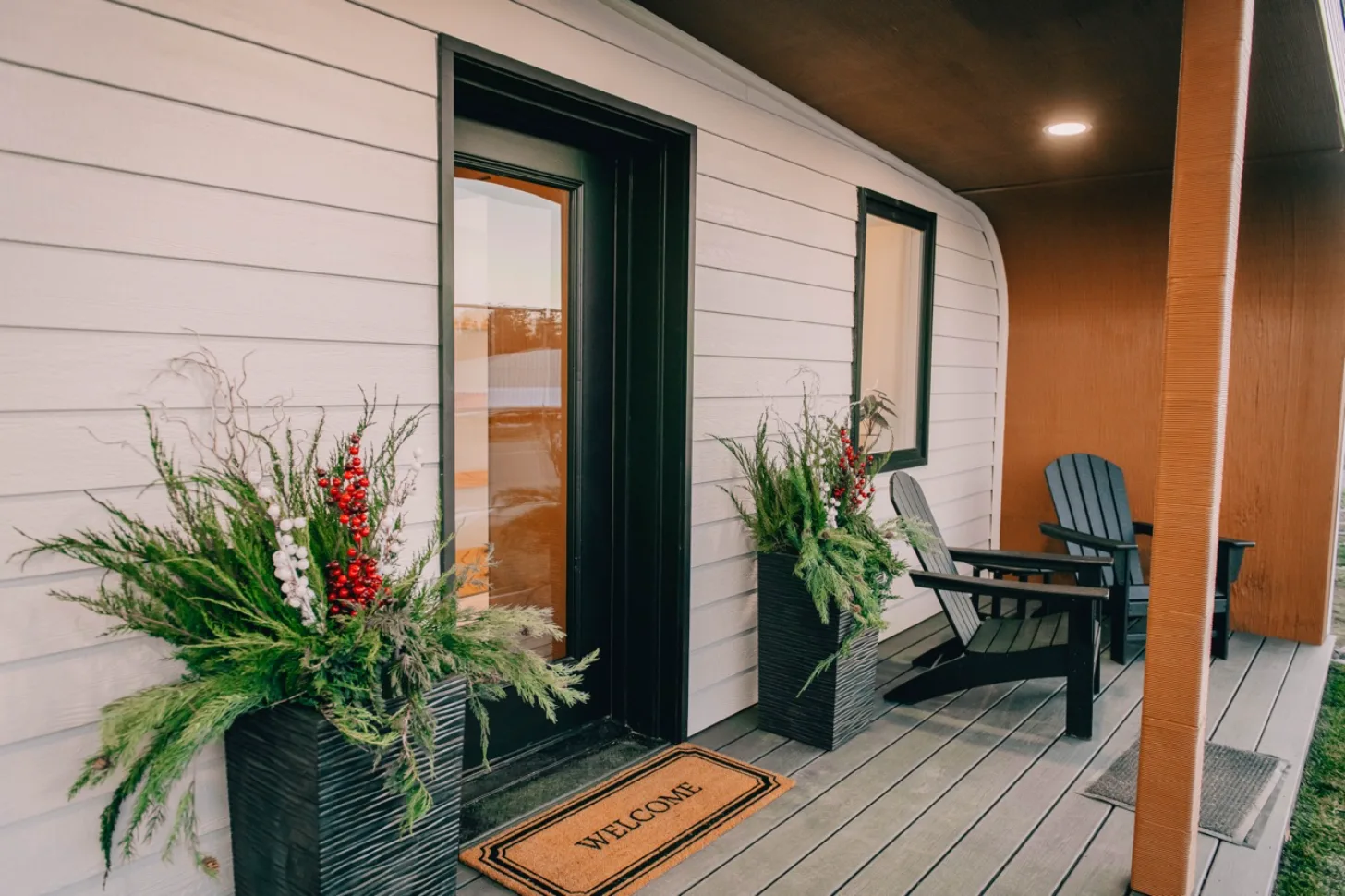 A modern porch entrance with white horizontal siding features a black door with a window, flanked by two black Adirondack chairs. Potted plants with red and white flowers sit on either side of the door. A "WELCOME" doormat lies on the gray composite decking. An orange brick wall is visible on the right side.