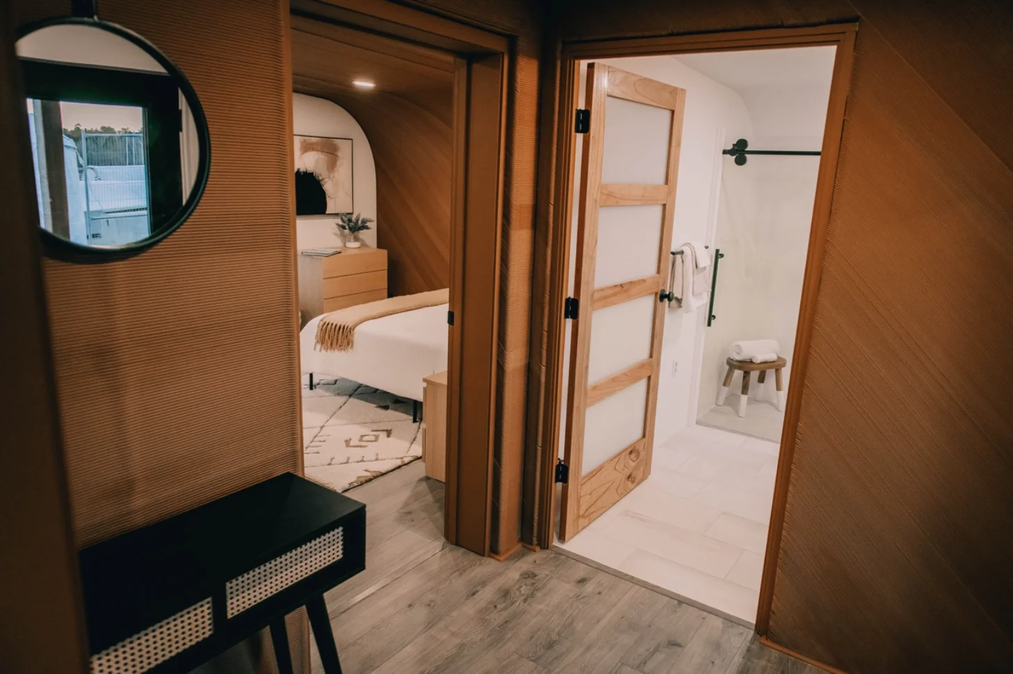 Interior view of a studio apartment showing a bedroom with white bedding and wooden furniture, an open bathroom doorway with a small stool, and a vanity area with a round mirror on the left. Wood-framed glass doors separate the rooms.