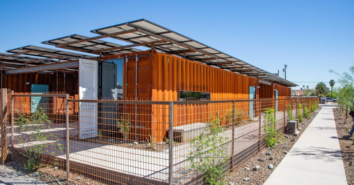 Row of orange shipping containers converted into residential units with solar panel canopies overhead, metal fencing, concrete walkway, and desert landscaping under clear blue sky.