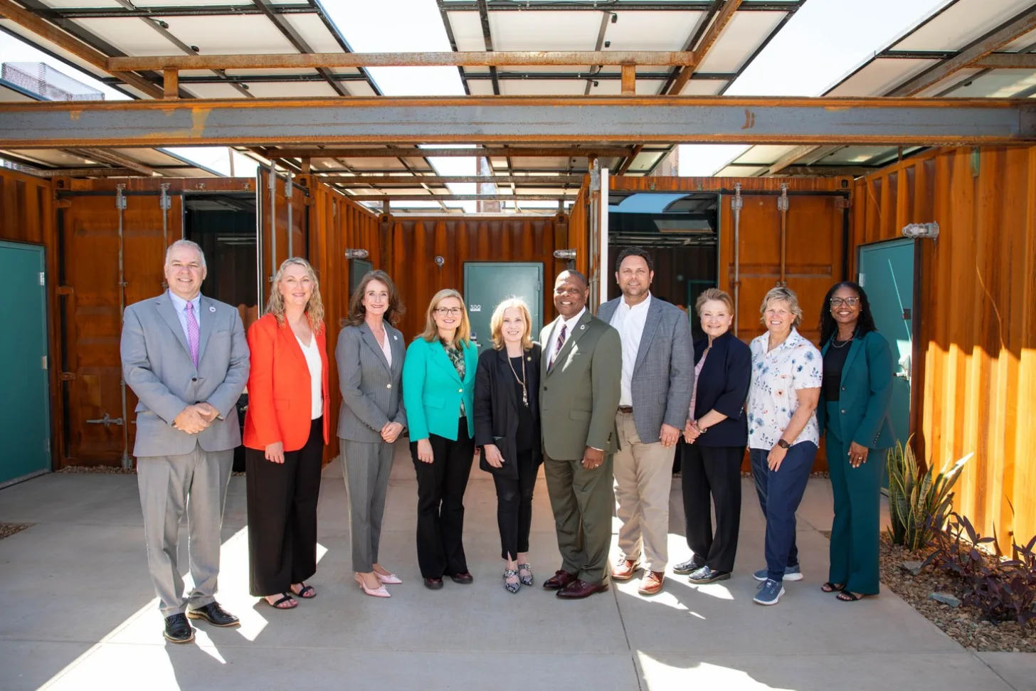 Ten people stand in a line inside a structure with orange shipping containers and a metal framework roof with skylights.