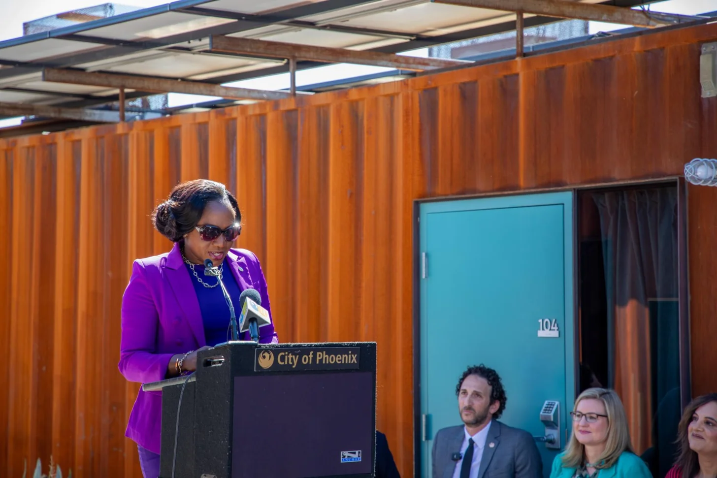 Woman in purple blazer, Phoenix Vice Mayor Kesha Hodge Washington, speaking at podium labeled "City of Phoenix" while three people seated nearby listen.