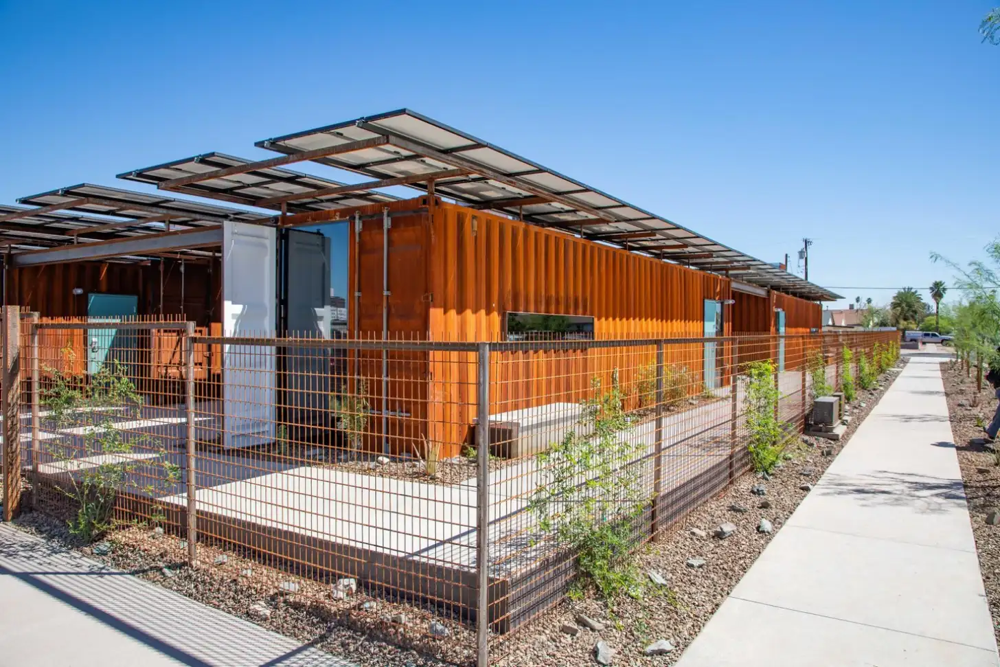 Row of orange shipping container structures with solar panel canopies, metal fencing, and concrete walkway on a clear day.