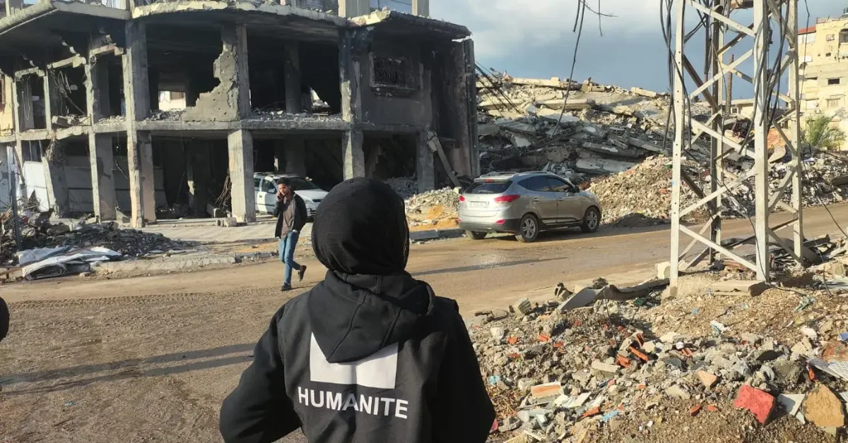 Person wearing a black jacket with "HUMANITE" written on the back stands in foreground, viewing destroyed concrete buildings and rubble in an urban area.