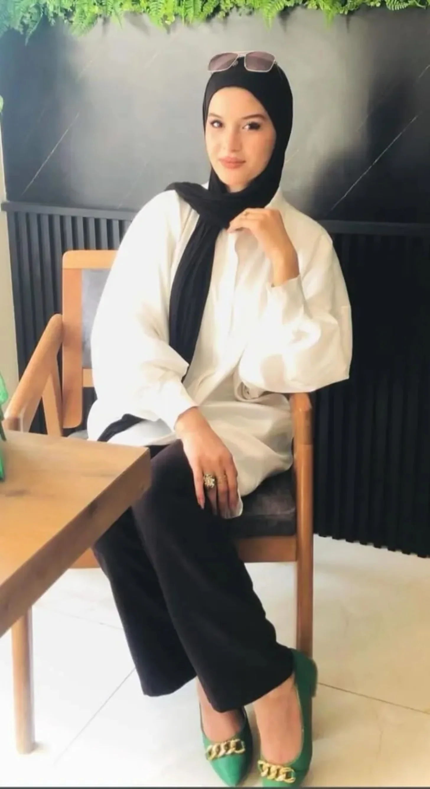 Woman seated on a wooden chair wearing a white blazer, black hijab, black cropped pants, and green shoes with gold chain detailing. She has sunglasses pushed up on her head and is resting one arm on her leg.