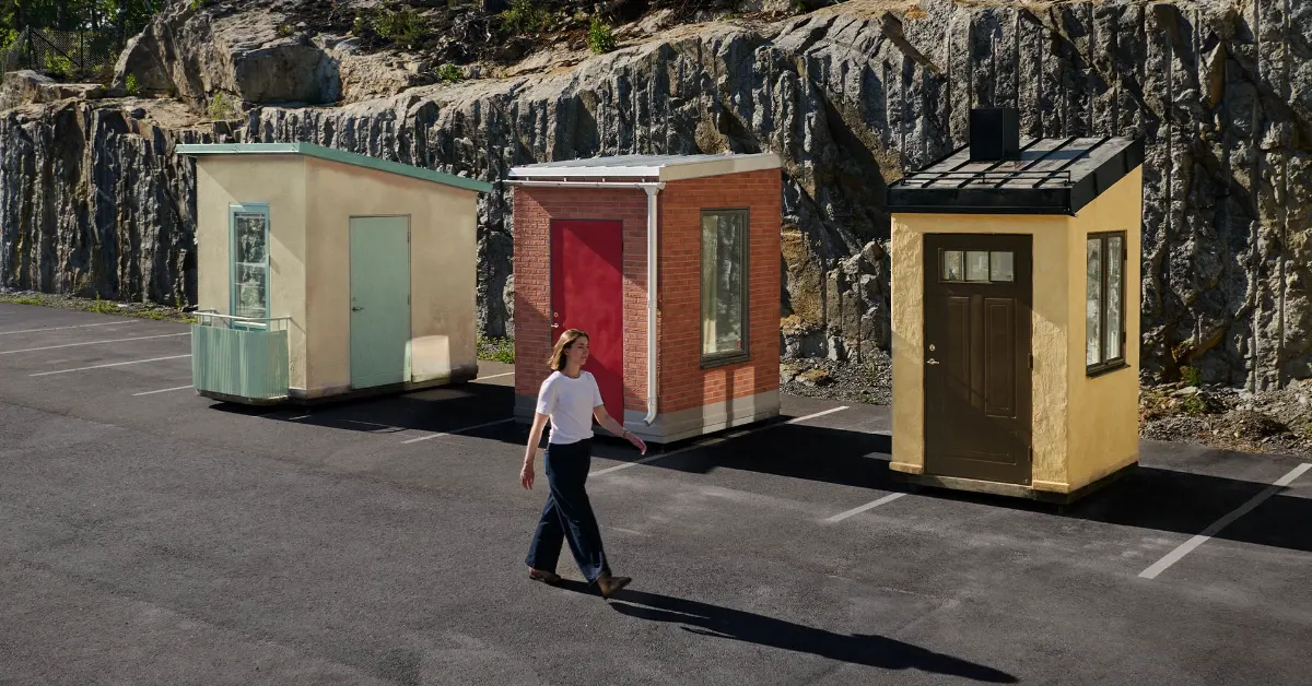 A woman in a white shirt and dark pants walks across a paved area in front of three small colorful structures—one tan with a green-trimmed door, one red brick with a red door, and one yellow with a brown door—set against a backdrop of tall, columnar rock formations.