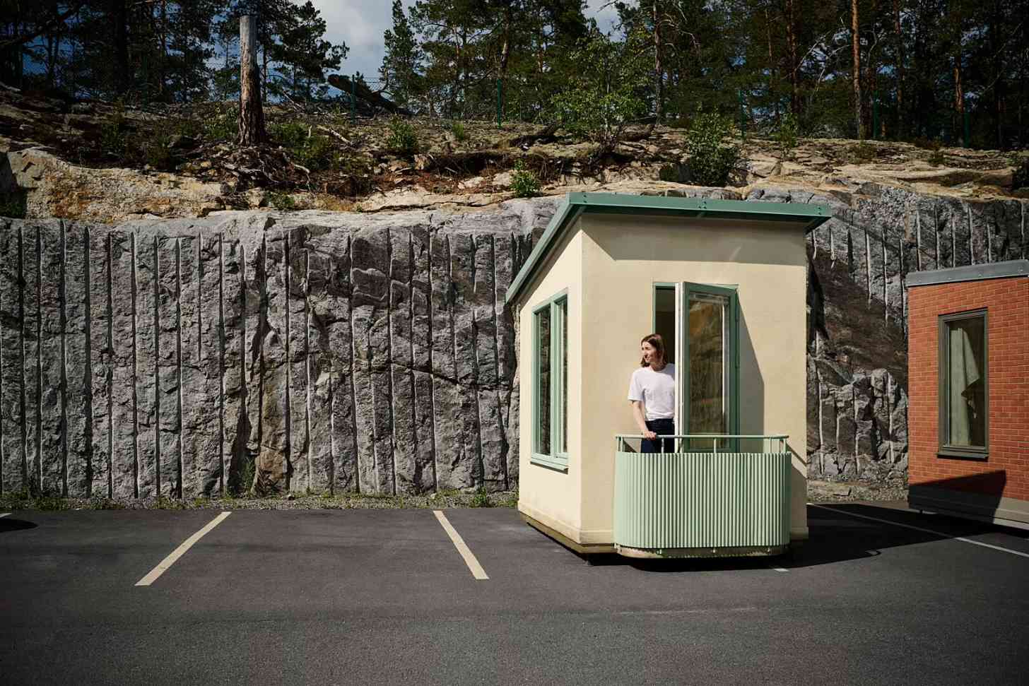 A woman stands on a small cream-colored building with a green railing and balcony, positioned in front of a tall gray stone wall in a parking lot with wooded hillside behind.