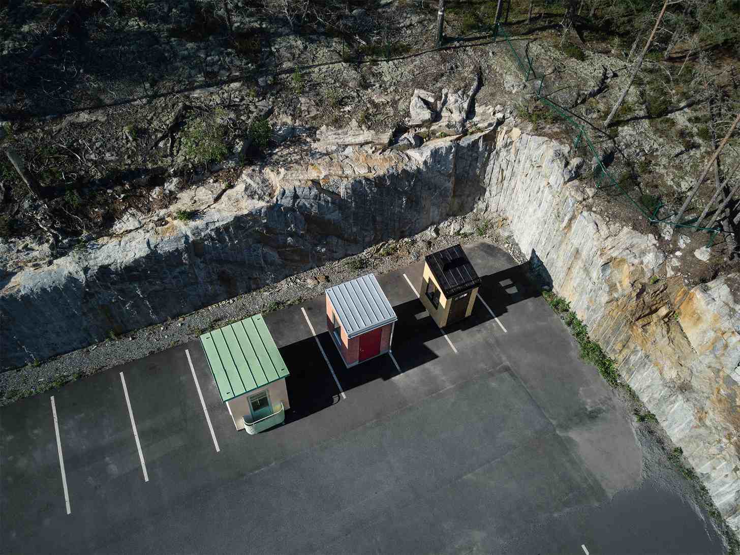 Aerial view of three small colored structures with metal roofs parked in a paved lot next to a quarry with steep white and gray stone walls.