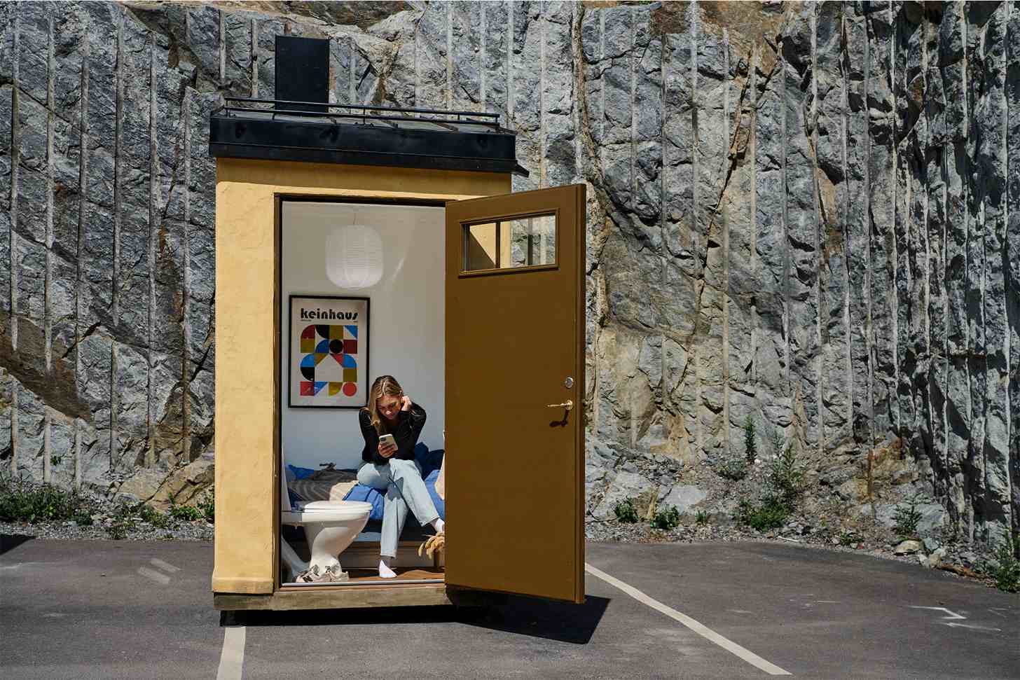 A woman sits inside a small yellow structure with a brown door, positioned against a large stone wall. A "keinohaus" poster is visible on the interior wall.
