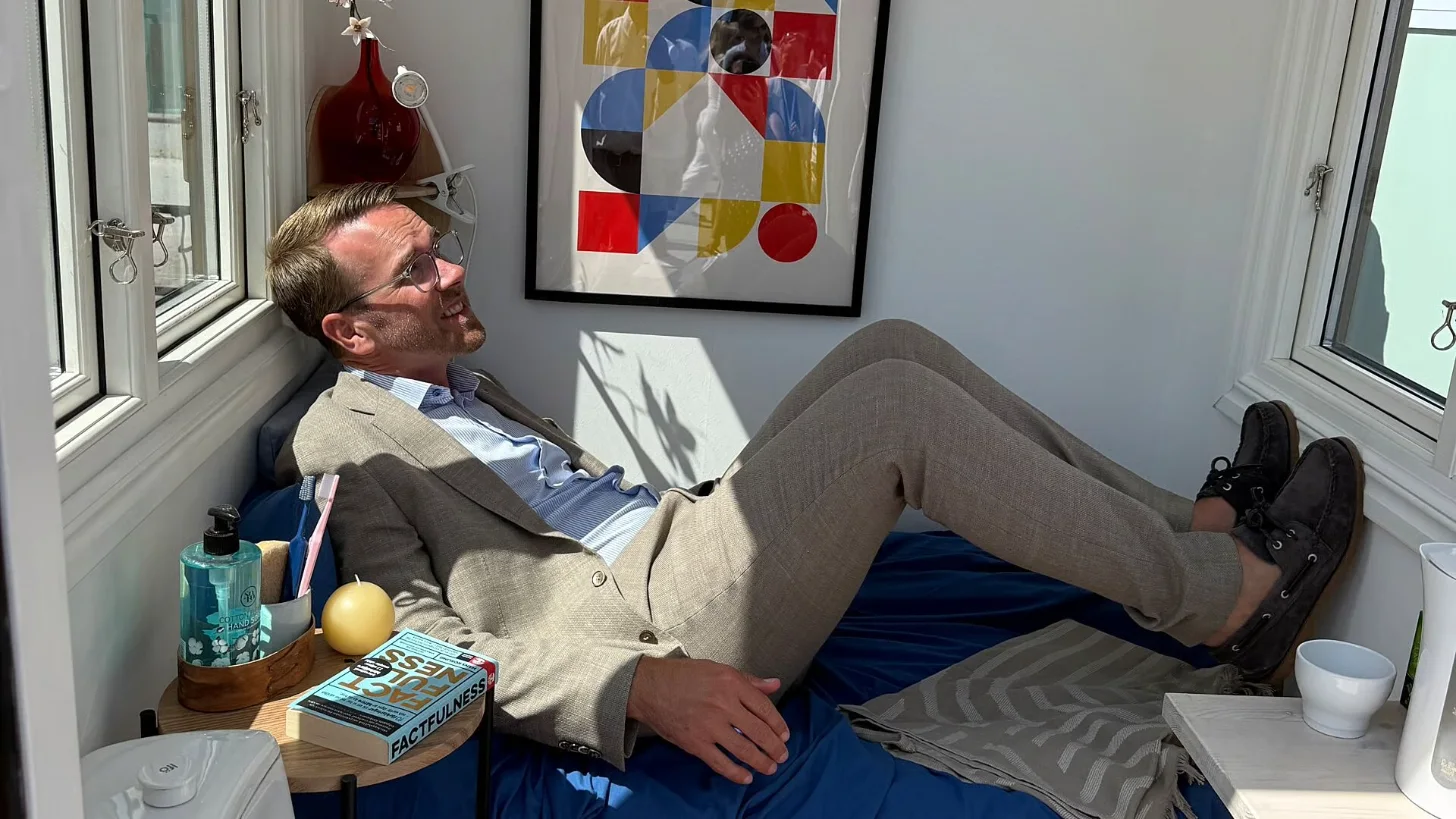 A man in a beige blazer and blue shirt reclines in a chair by a window, wearing sunglasses and black boots with feet up. A book titled "Peacefulness" sits on a small table beside him with a bottle of hand sanitizer and a yellow spherical candle.