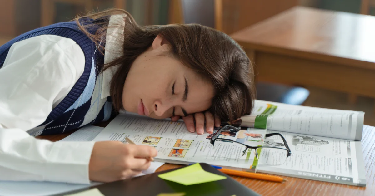 A student with dark hair rests her head on an open textbook at a desk, eyes closed, with glasses and a yellow sticky note nearby.