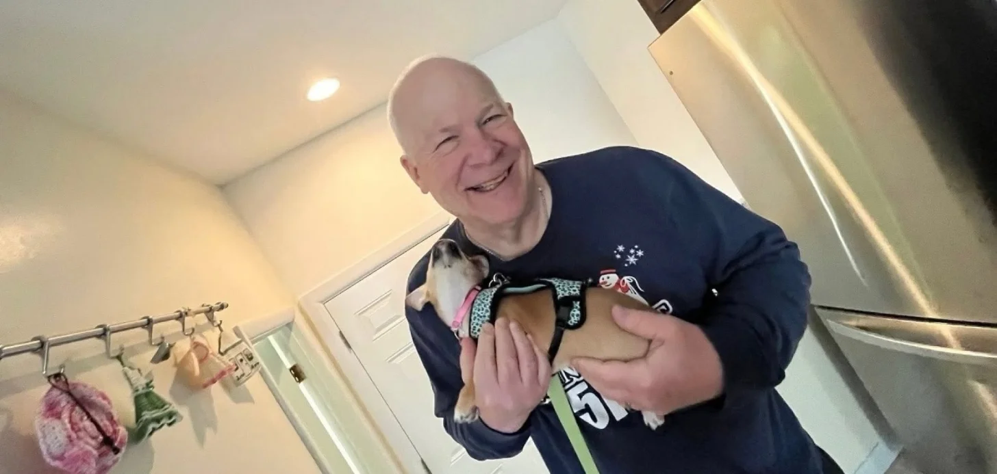 A man in a dark sweater with holiday decorations smiles while holding a small dog wearing a festive collar and leash indoors.