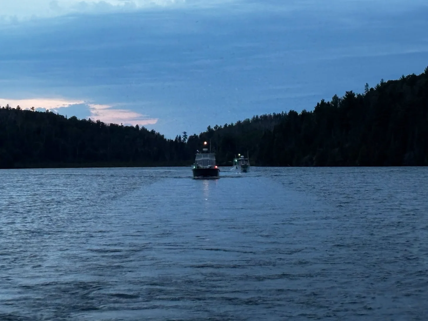 Two motorboats on a calm lake at dusk, with forested shoreline and evergreen trees silhouetted against a blue sky with clouds.