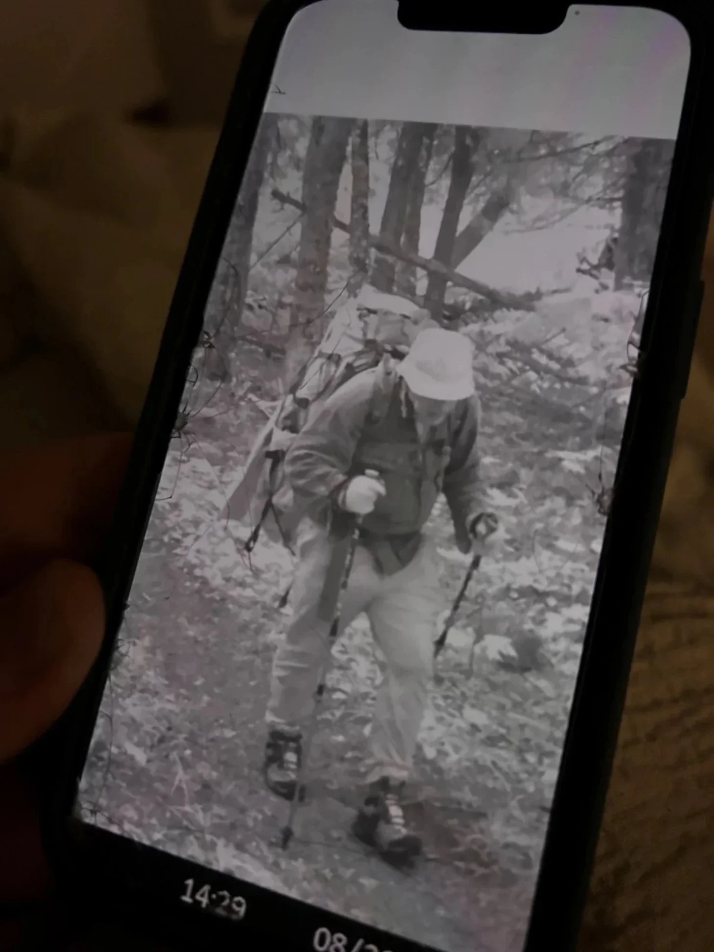 Black and white photograph displayed on a smartphone screen showing a person wearing a white hat and dark jacket standing on a wooded path with bare trees. Timestamp reads 14:29.