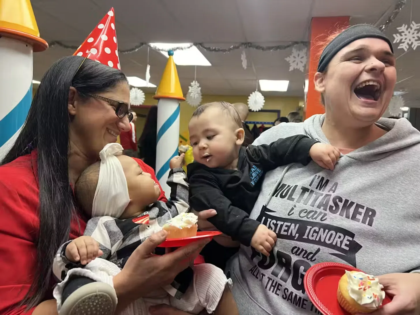 Two adults and two young children at a birthday party wearing paper party hats, holding cupcakes and smiling. Party decorations including garland and hanging snowflakes are visible in the background.