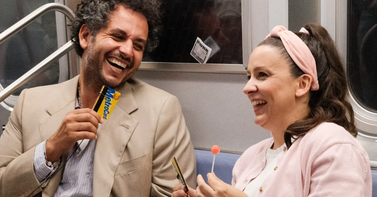 Two people on public transit laugh. The man on the left wears a beige blazer and holds a yellow and blue Metrocard. The woman on the right wears a pink headband and light pink cardigan, holding a lollipop.