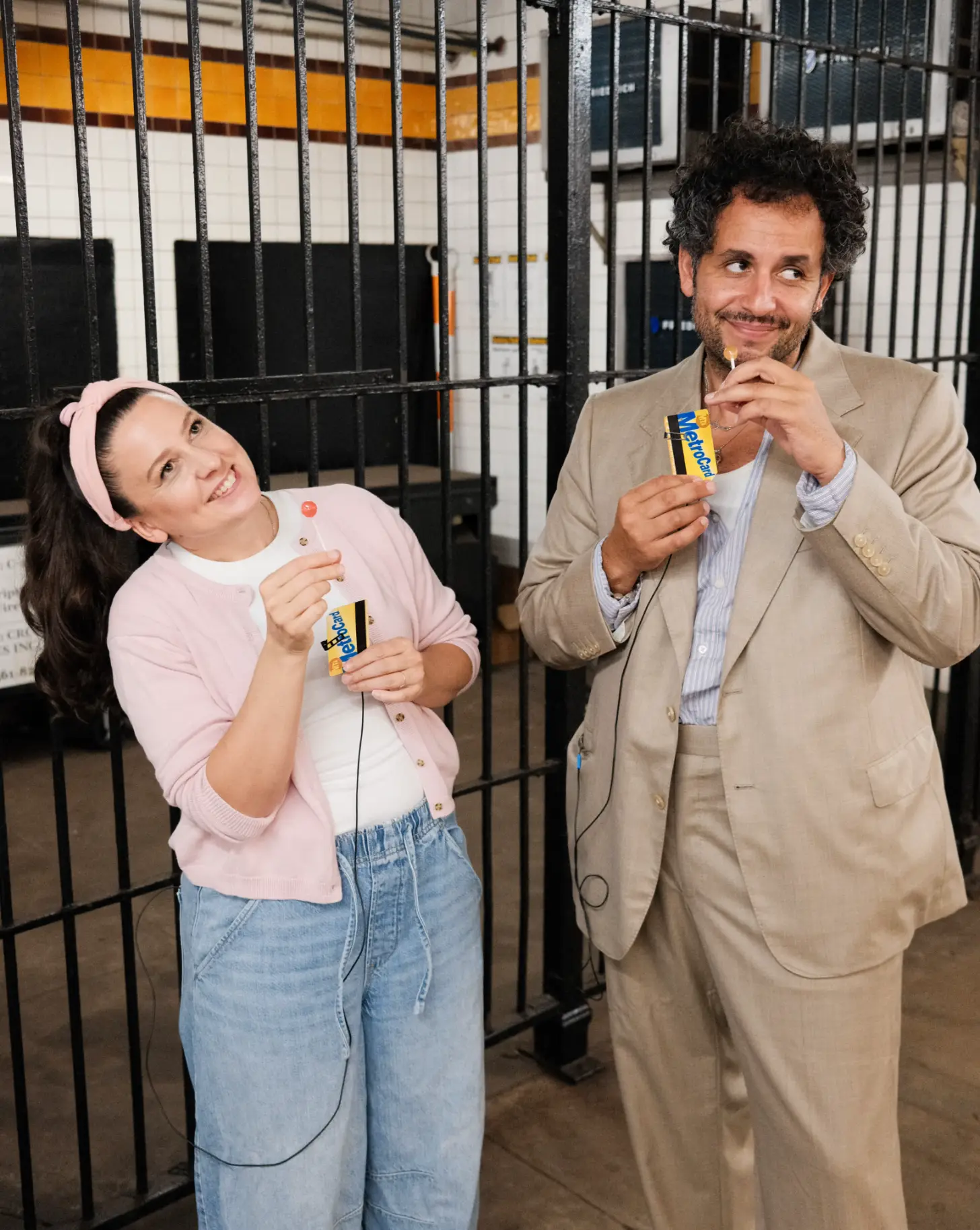 Two people holding Metrocards stand in front of black metal walls in a subway station. Both are smiling.