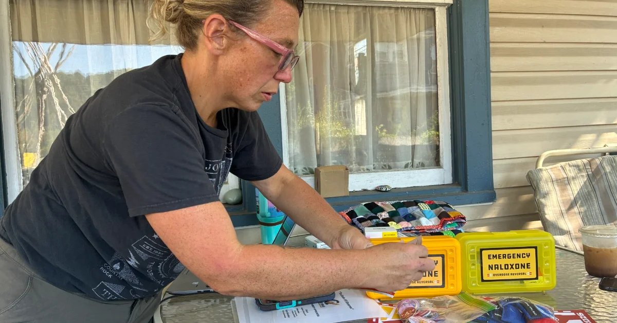 Woman wearing glasses and a dark shirt holds yellow and green boxes labeled "Emergency Naloxone" at a table with harm reduction materials and educational pamphlets visible.