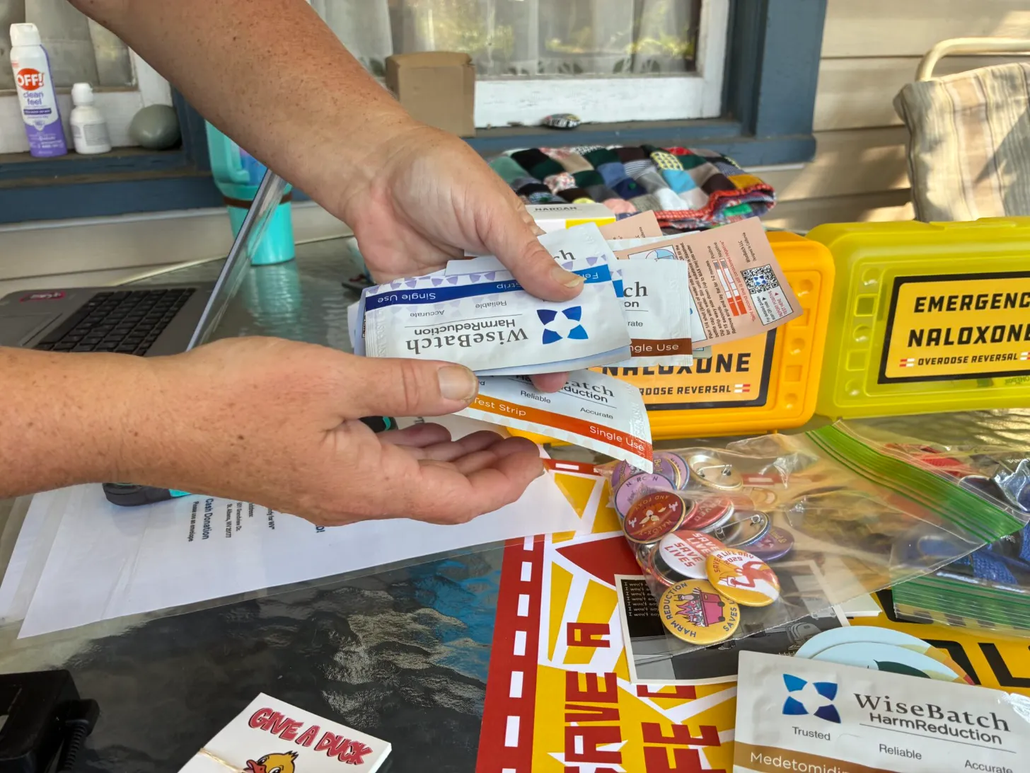 A person holds several medication boxes and test strips in their hands, including a yellow Narcan nasal spray box and packages labeled "WiseBatch HarmReduction" and "Medetomidine." A desk behind them displays various harm reduction supplies, including a yellow emergency naloxone kit, colorful bracelets, and scattered papers and materials.