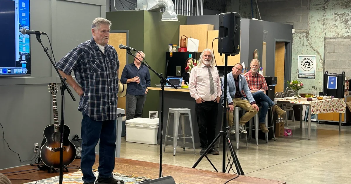 A man in a plaid shirt speaks at a microphone while holding a guitar in an indoor venue. Four other people sit on stools behind him, listening.