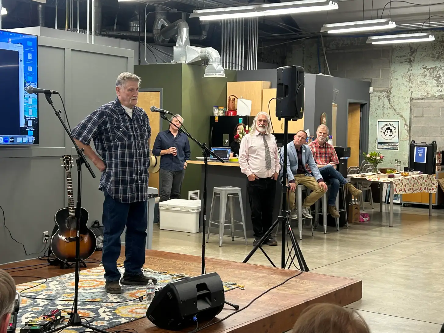 A man in a plaid shirt stands at a microphone holding a guitar, speaking to an audience seated on stools in an industrial workshop space with green walls and fluorescent lighting.