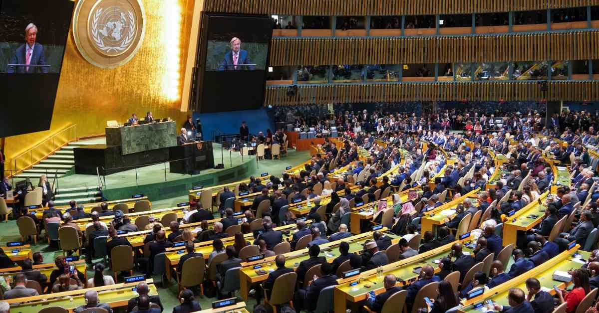 A speaker addresses the United Nations General Assembly from the podium as hundreds of delegates seated at desks listen. The UN emblem is visible on the wall behind the rostrum, with video screens displaying the speaker above.