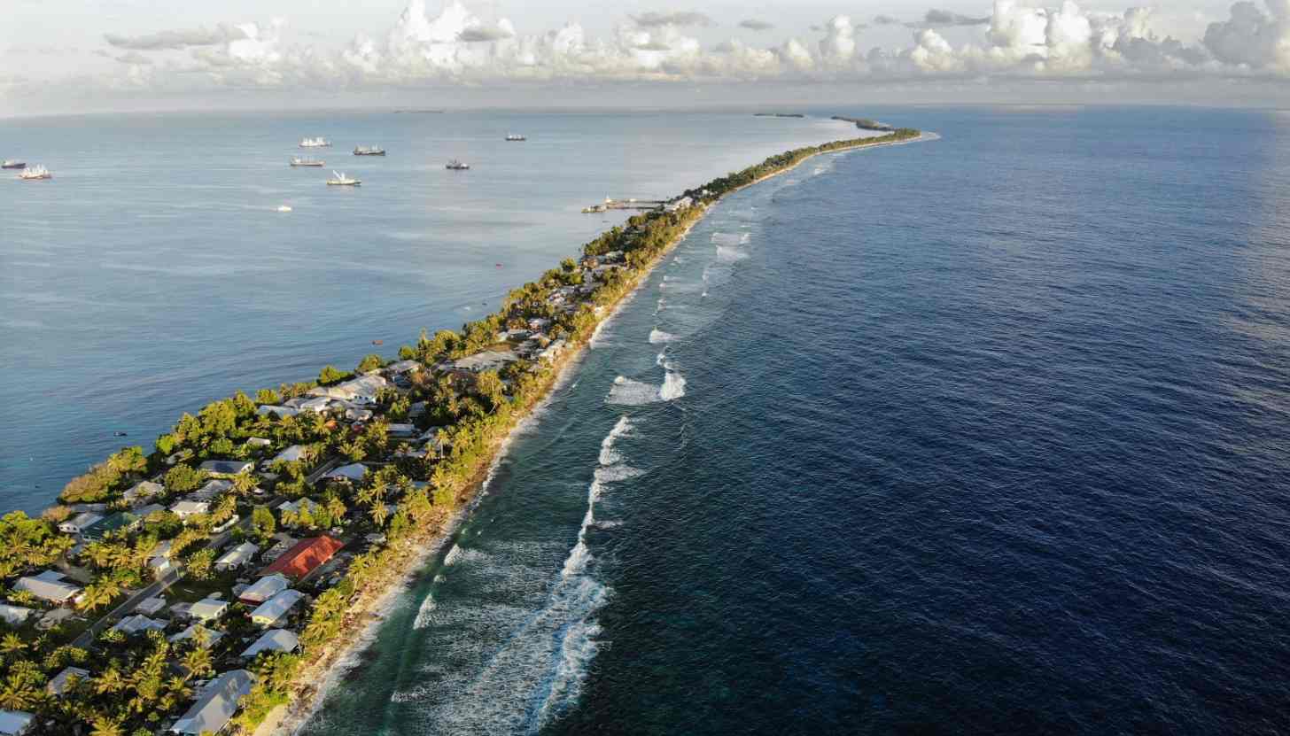 Aerial view of a narrow, curved spit of land lined with buildings and palm trees, separating a shallow lagoon with anchored boats from deeper ocean waters with white-capped waves.