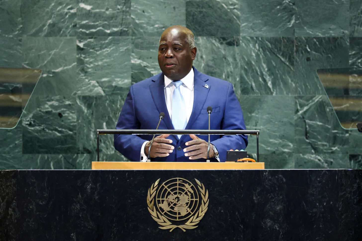A man in a blue suit and light blue tie stands at a United Nations podium, speaking with his hands positioned on the lectern. The UN emblem is visible on the front of the podium, with a green marble wall behind him.