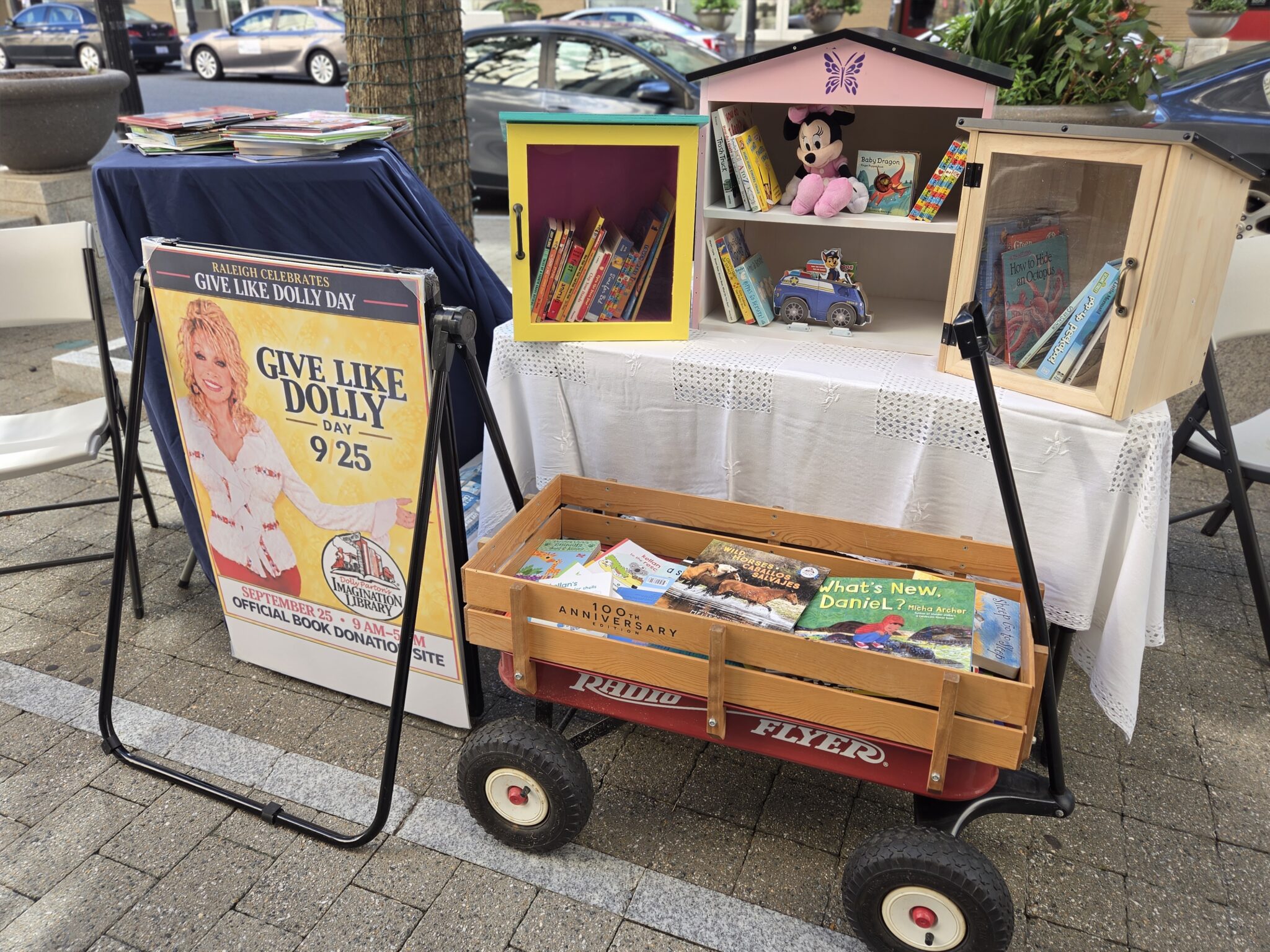A cart full of books beneath various tables with more books atop them, beside a sign that says: "Give Like Dolly" with Dolly Parton smiling on it