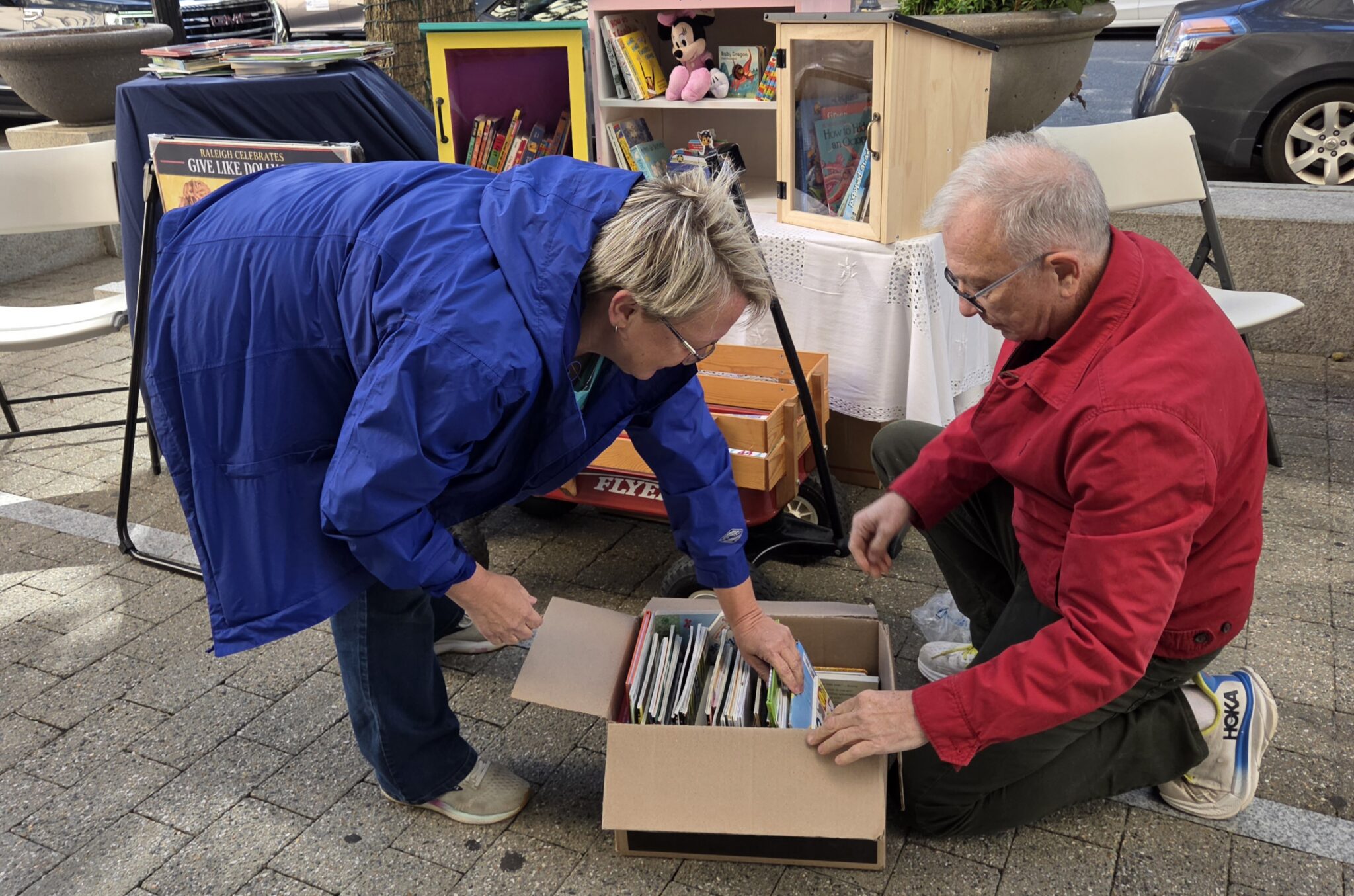 A woman in a blue jacket bends down to leaf through a box of books as a man in a red jacket kneels on the ground beside it