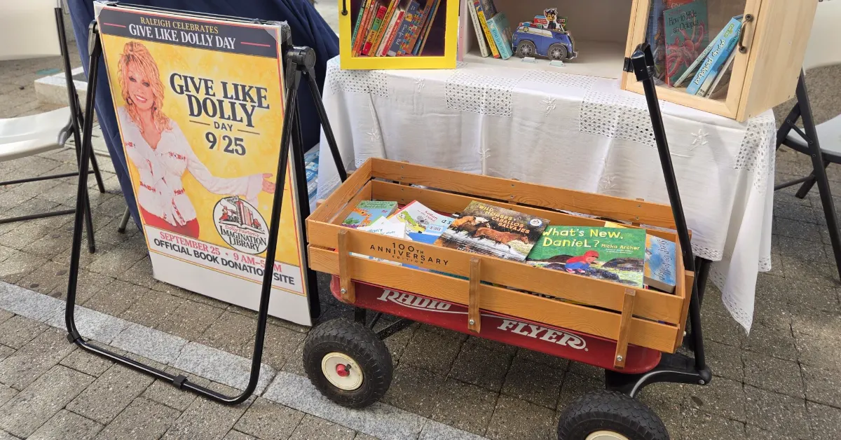 A cart full of books beneath various tables with more books atop them, beside a sign that says: "Give Like Dolly" with Dolly Parton smiling on it