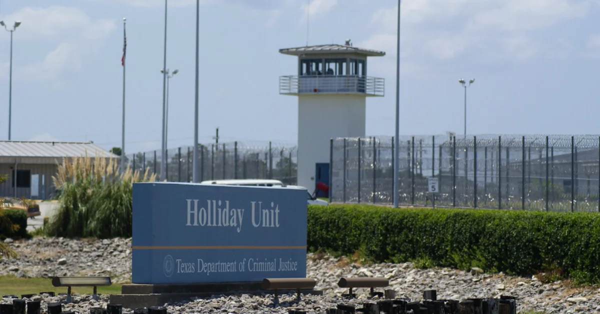 A blue sign reading "Holliday Unit" and "Texas Department of Criminal Justice" stands in front of a prison facility with a tall guard tower, chain-link fencing topped with barbed wire, and perimeter lighting.