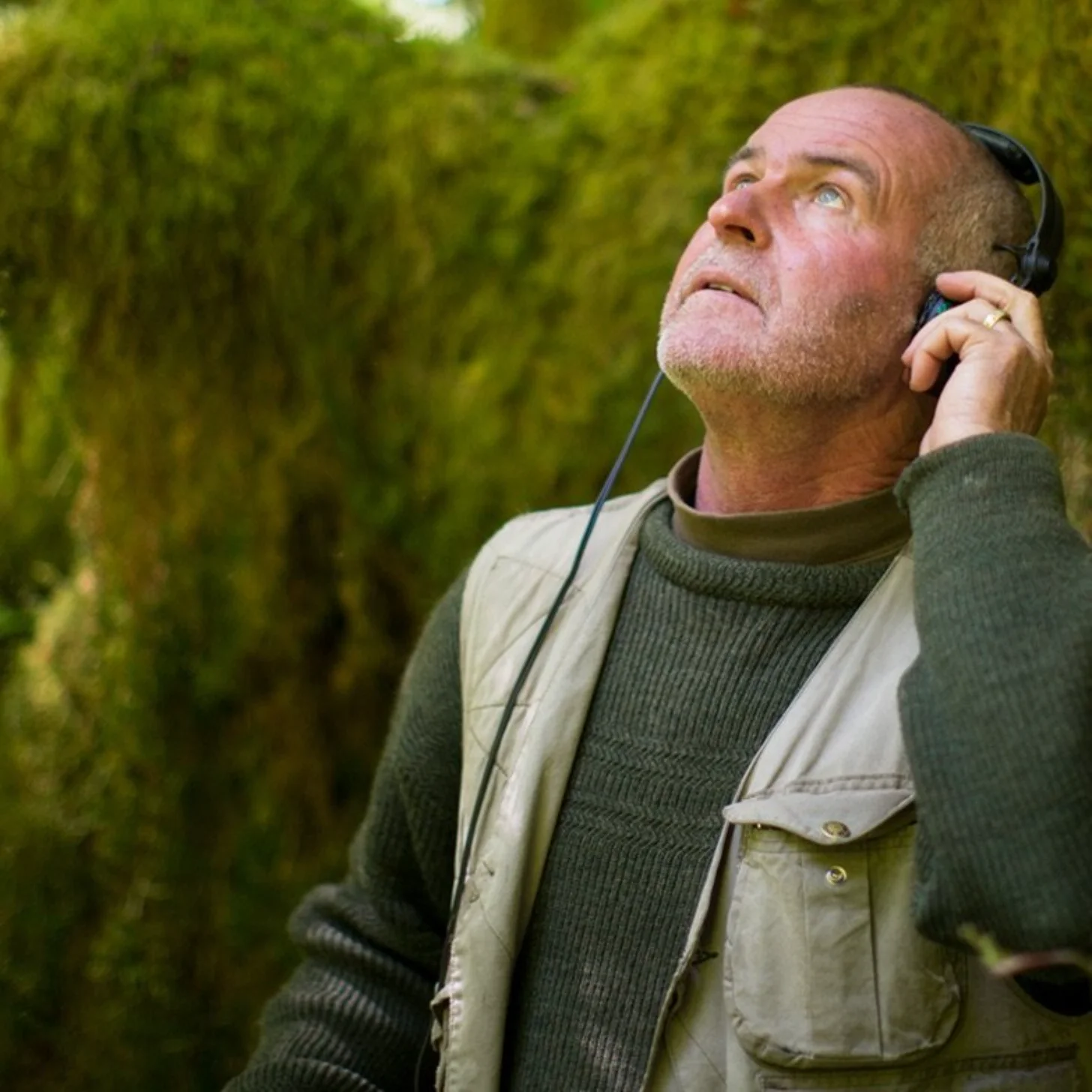 A bald man wearing a olive-green turtleneck and tan vest holds headphones to his ear while looking upward outdoors, with blurred green foliage in the background.