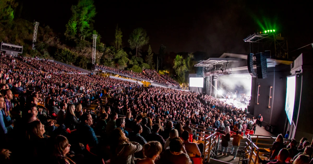 Thousands of people fill an outdoor stadium seating area at night, facing a large illuminated stage with screens visible on the right side. Tall green-lit towers and trees frame the venue.