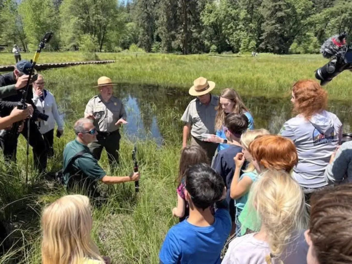 Park ranger in tan uniform and hat speaks to a gathered group of visitors in a grassy meadow with a shallow stream and forested hillside in the background. Camera operators film the presentation from both sides.