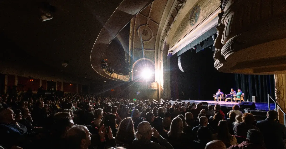 A large audience fills an ornate theater with a curved balcony while three hosts from The Bulwark sit on stage under bright lighting.